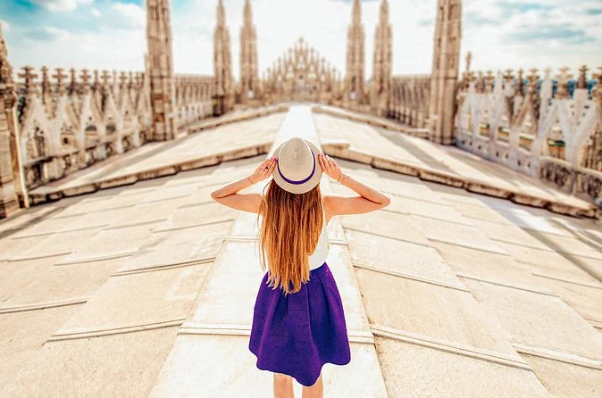 A woman in a white hat and purple skirt seen from behind on the sloping marble roof of the Duomo, with spires ahead