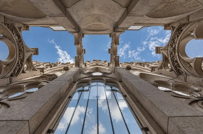 Looking straight up through a tall marble opening on the cathedral roof, with Gothic tracery framing blue sky and a large leaded window below