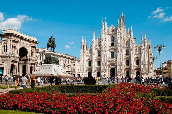 Piazza del Duomo with red flowerbeds in front, the equestrian statue and the cathedral facade behind under a blue sky