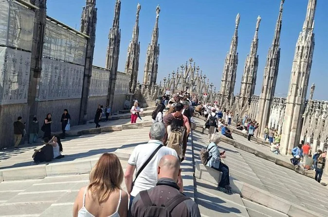 A dense crowd of visitors walking along the sloping central terrace of the cathedral roof on a sunny day, between spires and scaffolding