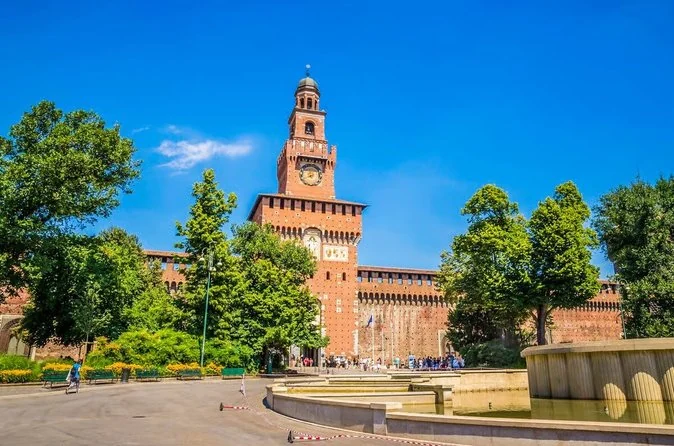 The red brick clock tower of Sforza Castle in Milan behind a round fountain and green trees on a sunny day