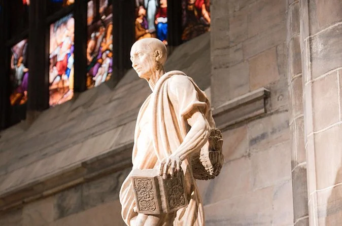 Marble statue of St Bartholomew inside Milan Cathedral, holding a book, with his flayed skin draped over his shoulders and stained glass behind