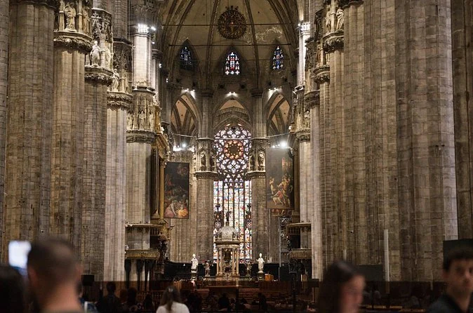The nave of Milan Cathedral with massive stone pillars leading to a tall stained glass window behind the high altar